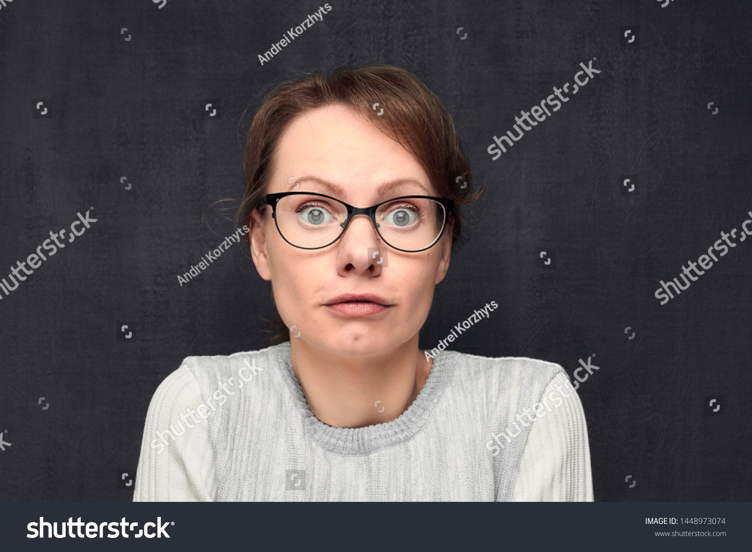 Studio close-up portrait of cute and funny caucasian fair-haired girl with eyeglasses, silly grimacing and looking naively and with bewilderment at camera, against gray background. Headshot #1448973074