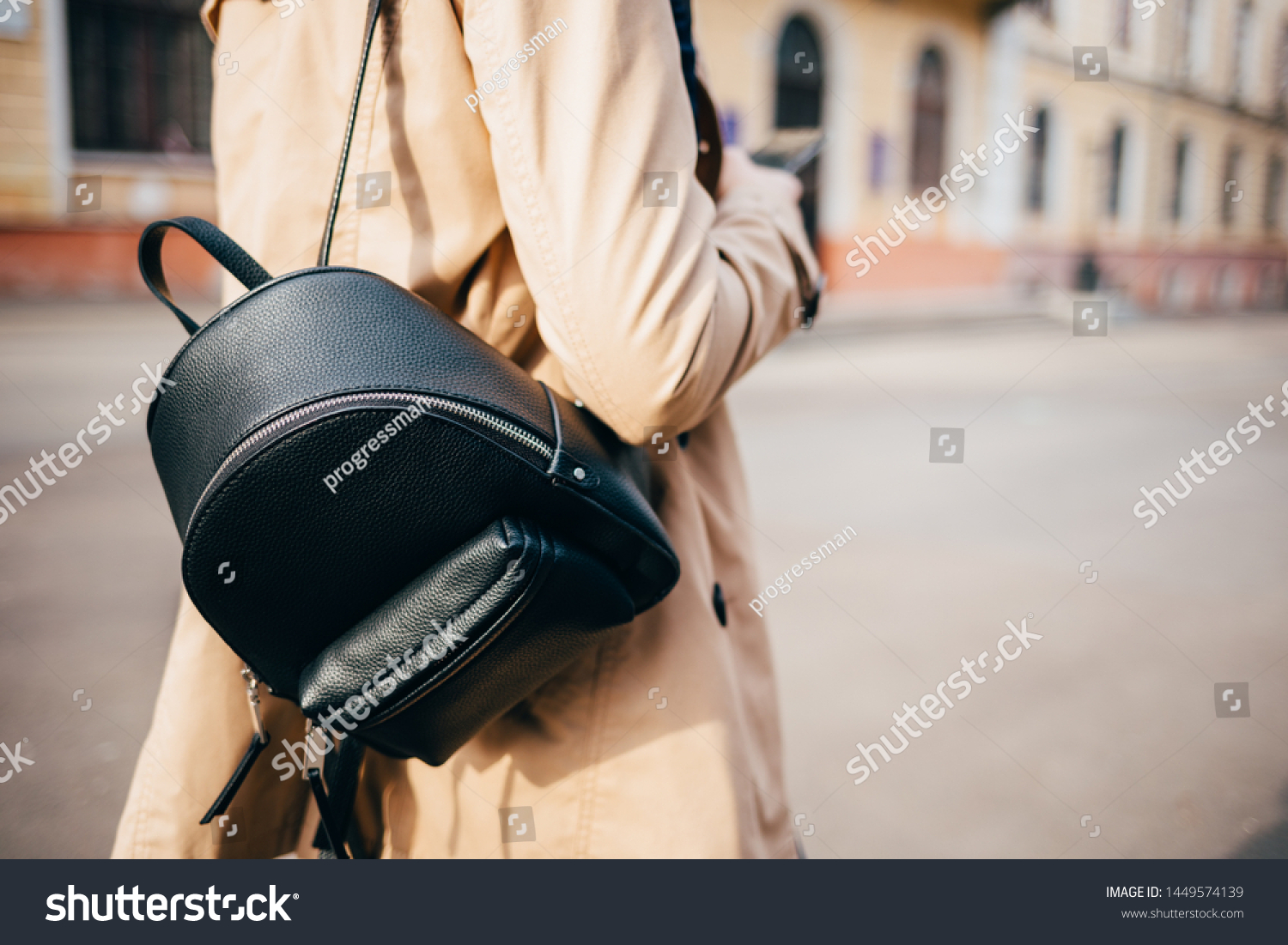 Close-up of elegant women's backpack. Rear view of young woman dressed in beige coat and with black bag walking down street in city. #1449574139