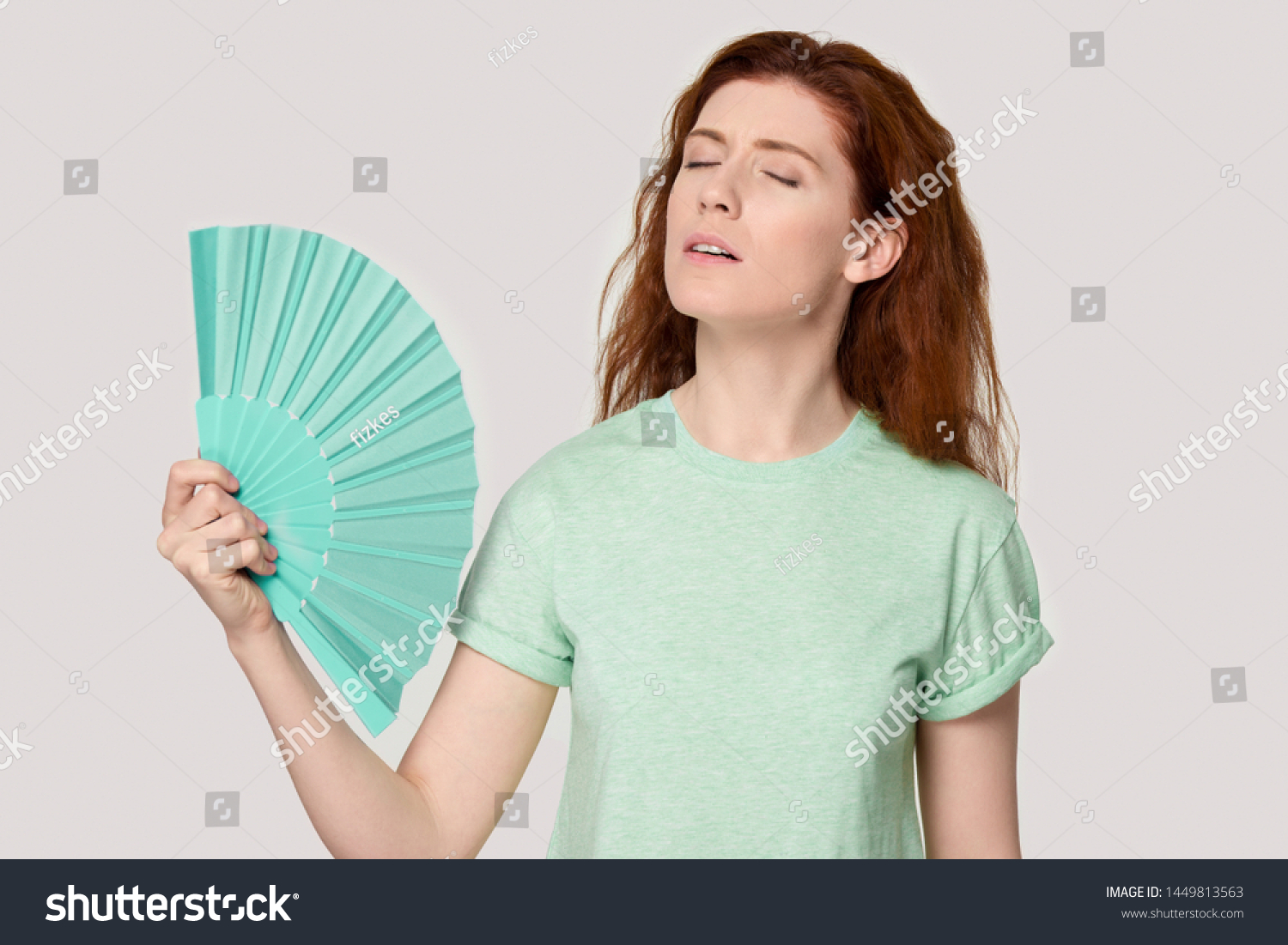 Overheated young red-haired woman using fan, cooling herself, suffering from hot summer weather or high temperature inside, heat stroke, feeling unwell, sweating, isolated on grey studio background. #1449813563