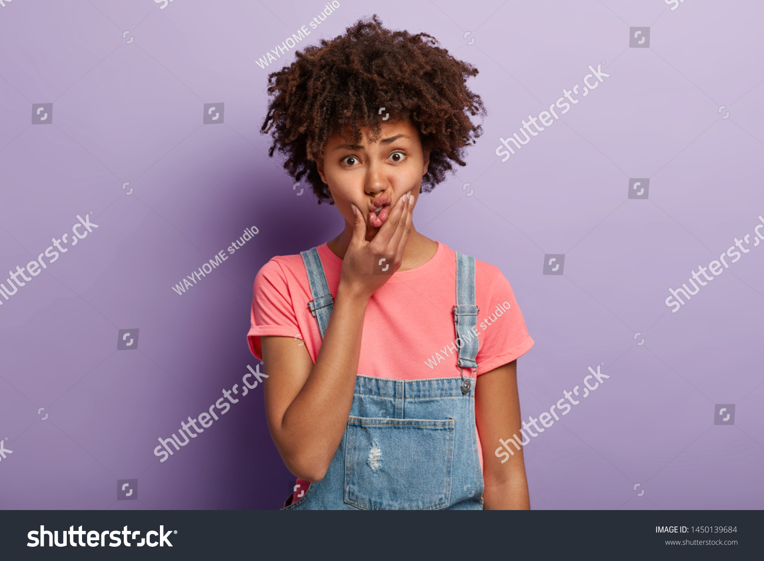 Discontent lovely lady presses lips with hand, makes grimace, has unhappy expression, thinks about something, dressed in stylish clothes, poses against purpe wall. Human facial expressions concept #1450139684