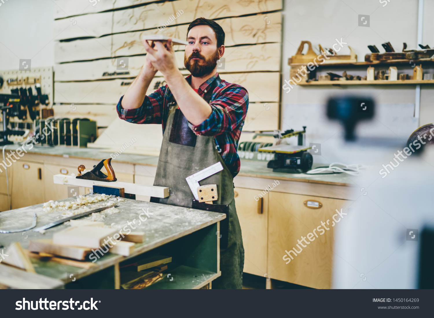 Bearded carpenter enjoying hobby working process at his workstation of manufacturing, side view of skilled self employed artisan in apron checking element for handmade construction of wooden plank #1450164269