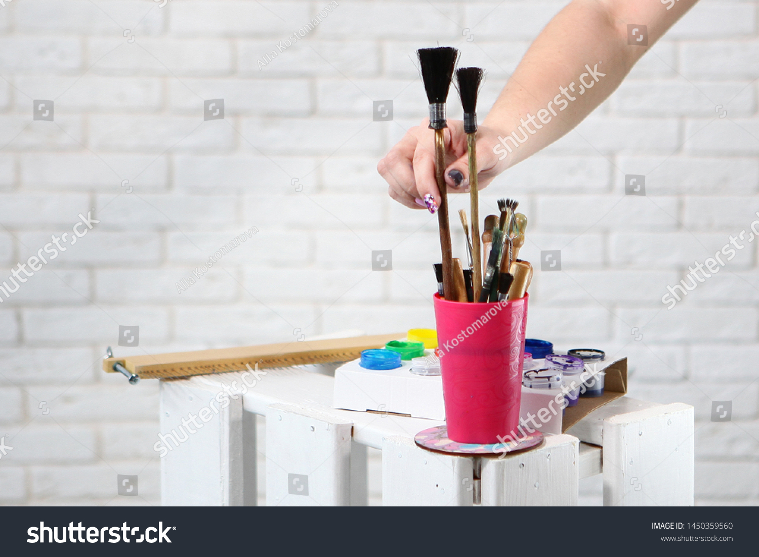 Female hand takes a brush for drawing. Brushes for painting are in the cup. Photo on the background of a white wall with a brickwork effect. Concept hobbies and hobbies. Photos in the interior. #1450359560