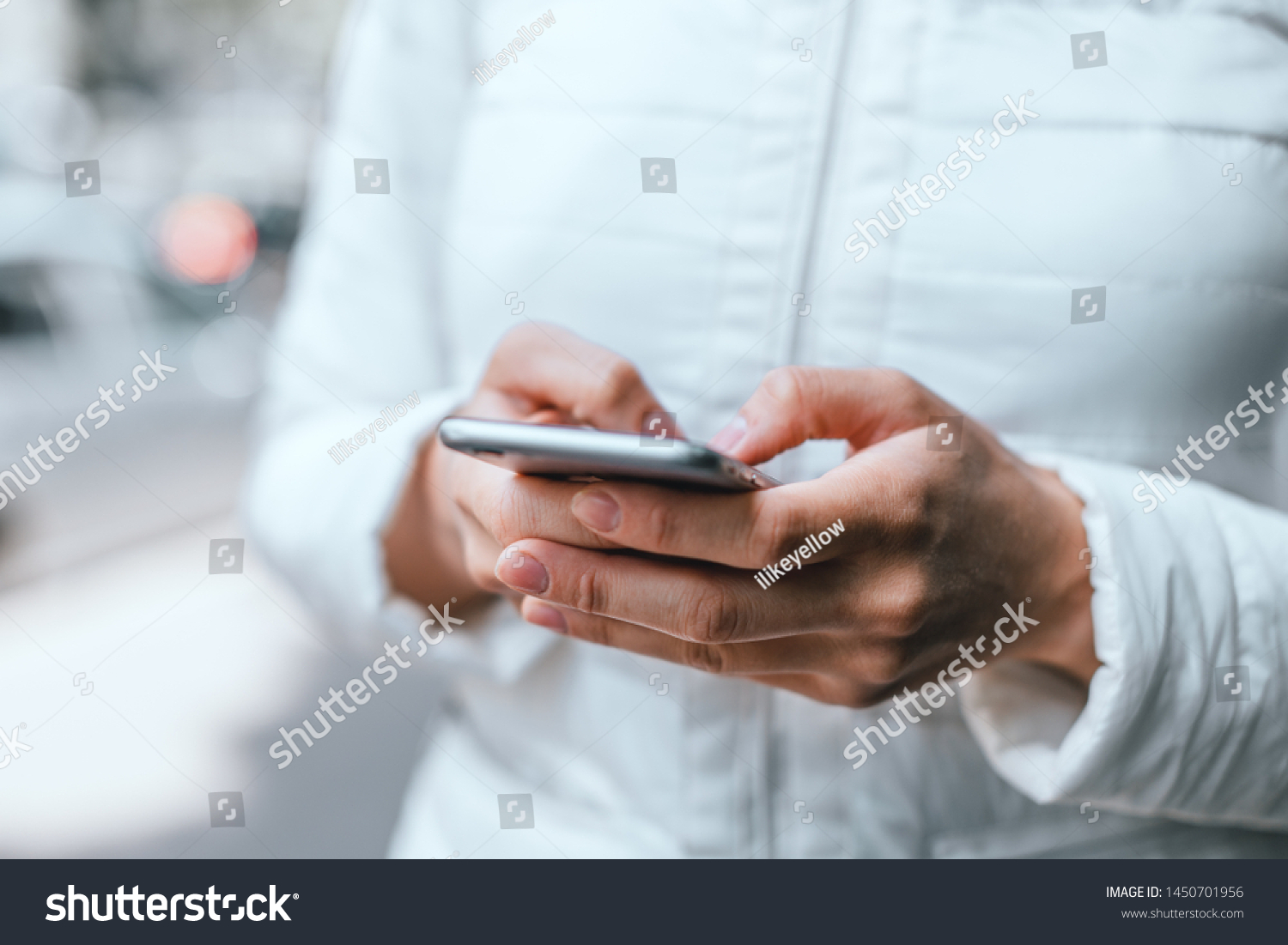 Close up of woman's hands in white jacket holding grey smartphone and typing a message with both hands on street. The side of the phone. #1450701956