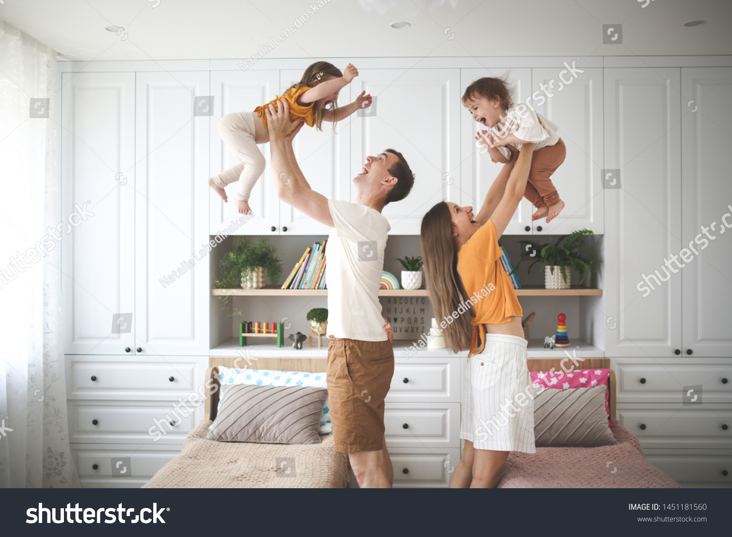 Beautiful Caucasian family with two children, dad and mom hold in their arms in the air two sisters toddlers, a child with Down syndrome in the family, a real interior #1451181560