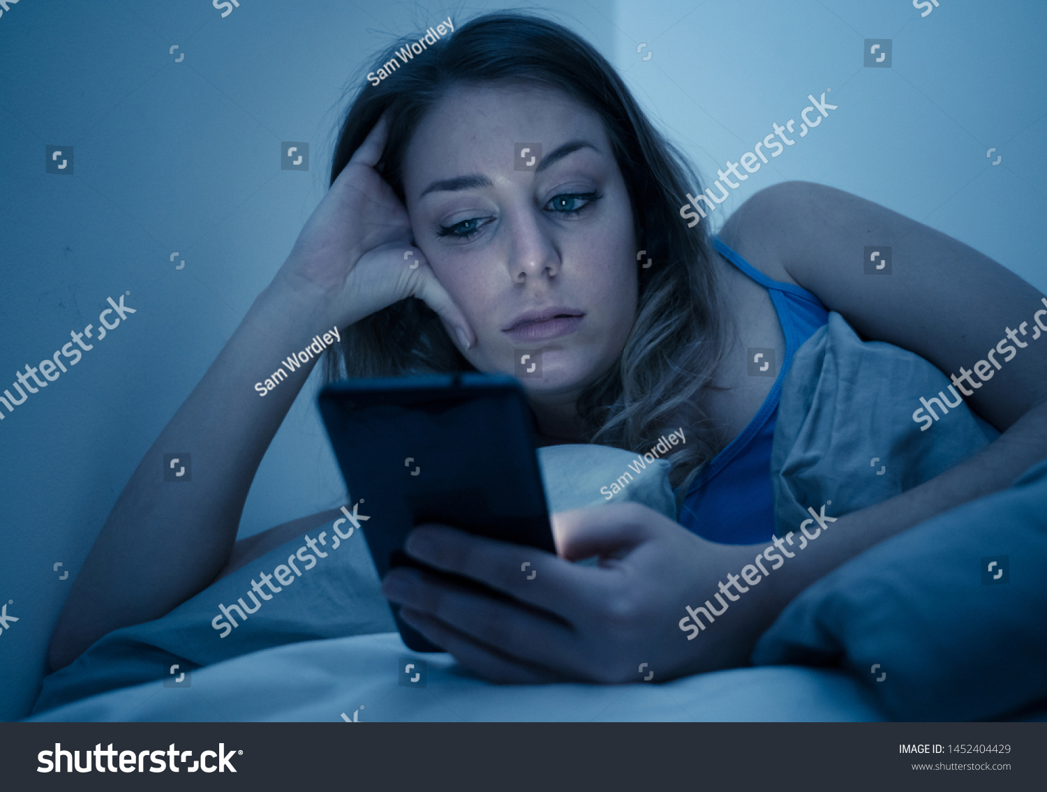 Addicted young beautiful woman in bed, chatting and surfing on the internet using her smart phone sleepy, dull and tired late at night in mobile addiction and technology overuse concept. #1452404429