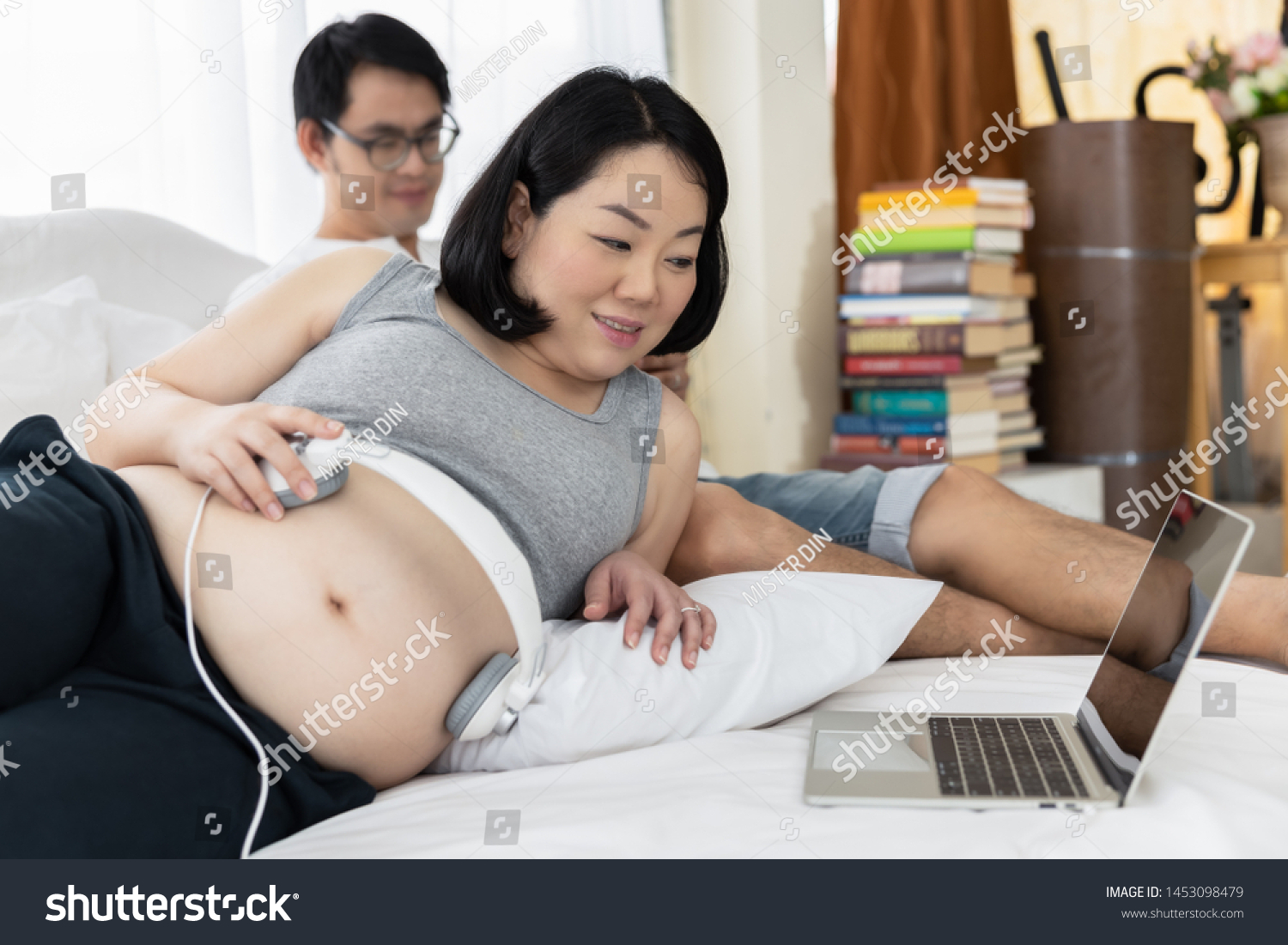 pregnant couples husband and wives resting on the bed in the bedroom and the wife opening the song on laptop let baby in the womb listen happy family baby love care concept #1453098479
