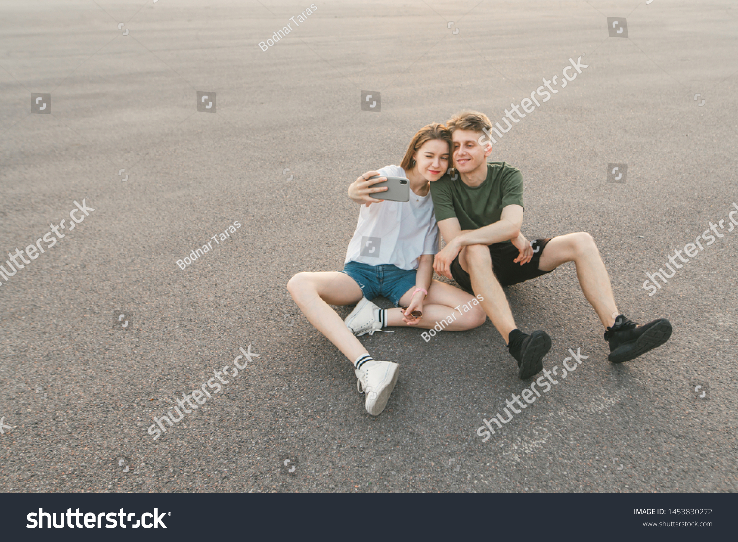 Funny couple loves sitting on the pavement and doing a sephi on a smartphone. Stylish young man and girl sit on the street and make a photo on the smartphone, posing to the camera with funny faces. #1453830272