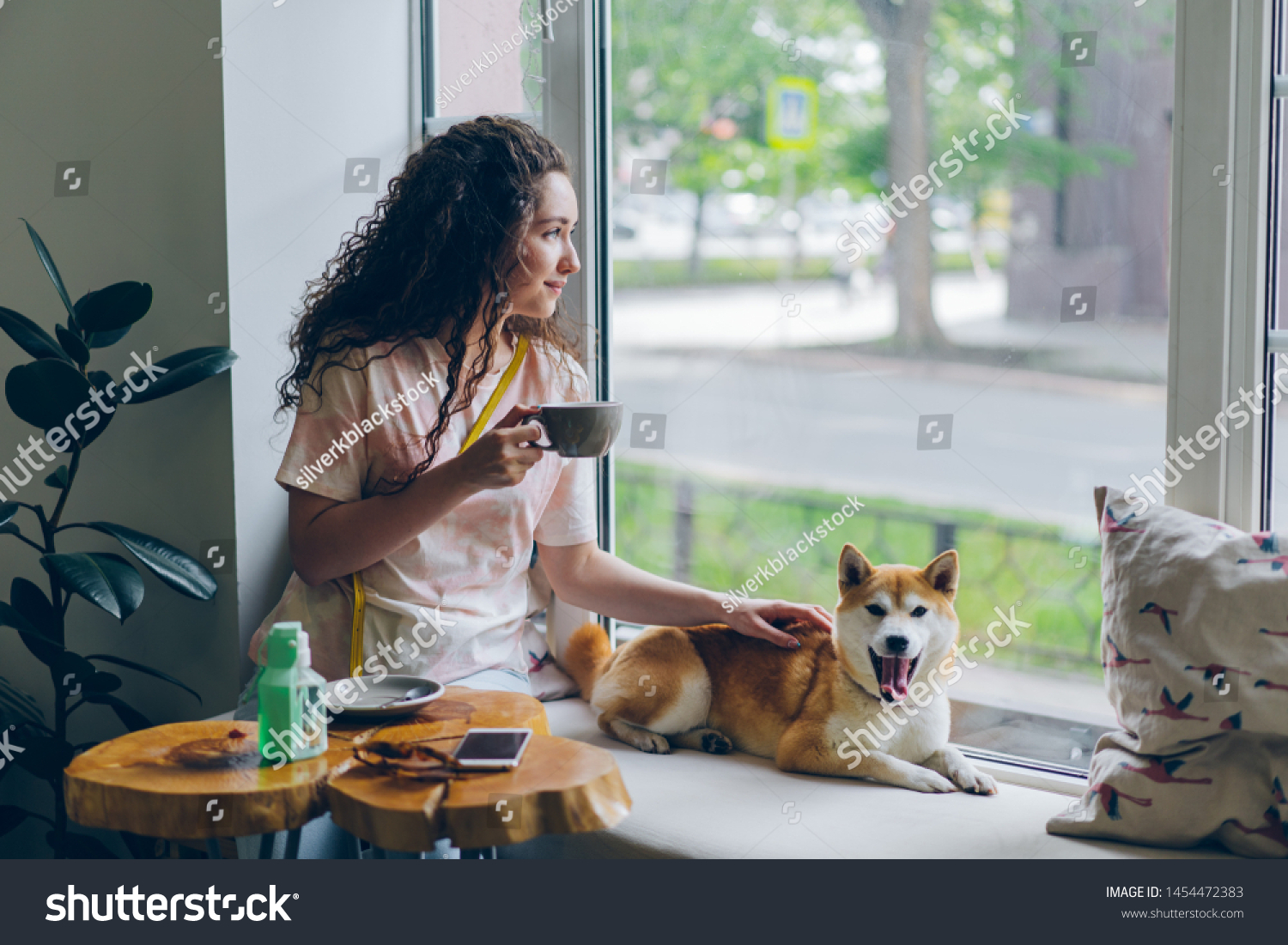 Happy student pretty girl is stroking adorable shiba inu dog sitting in cafe on window sill with cup of tea and enjoying leisure time. People and relaxation concept. #1454472383