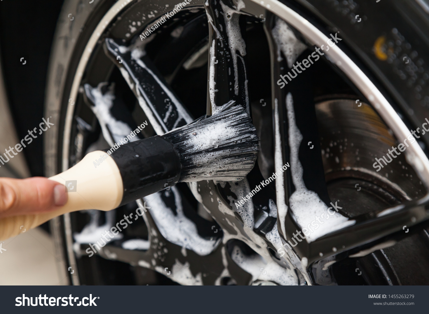 A male worker washes a black car with a special brush for cast wheels and scrubs the surface to shine in a vehicle detailing workshop. Auto service industry.