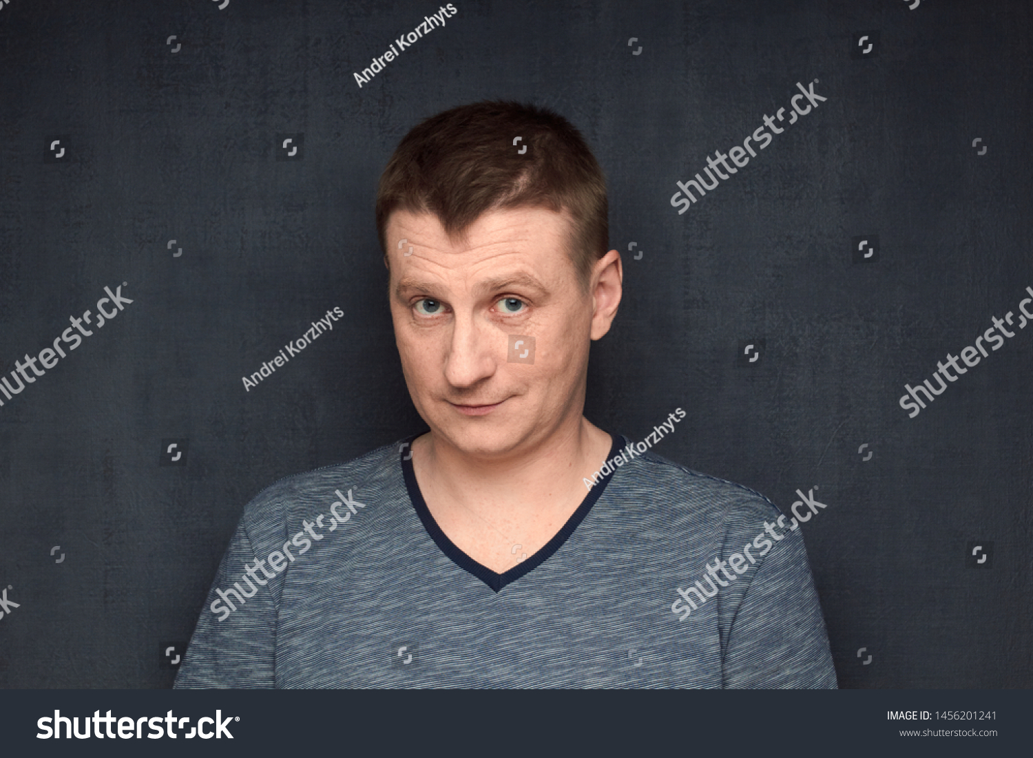 Studio close-up portrait of genial caucasian fair-haired man, wearing t-shirt, smiling kindly and and looking naively from under forehead at camera, making innocent face, against gray background #1456201241