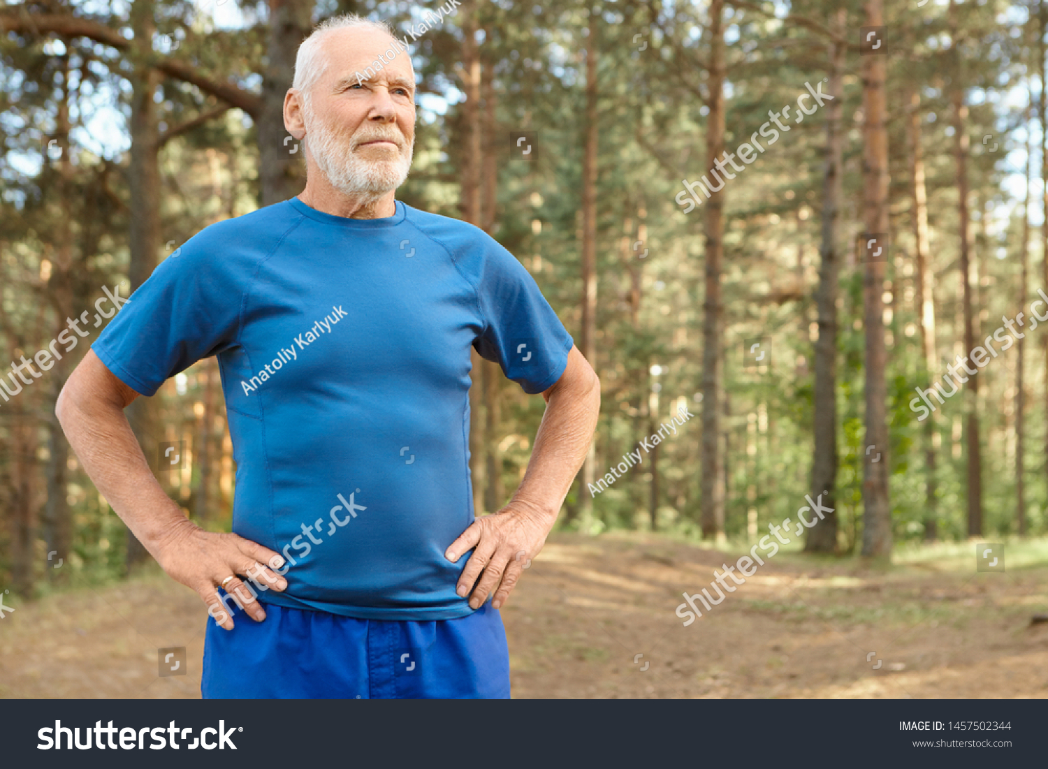 Self determined elderly man on retirement training outdoors in pine wood, holding hands on his waist, doing exercises to warm up body before run. Bearded retired male catching breath after workout #1457502344