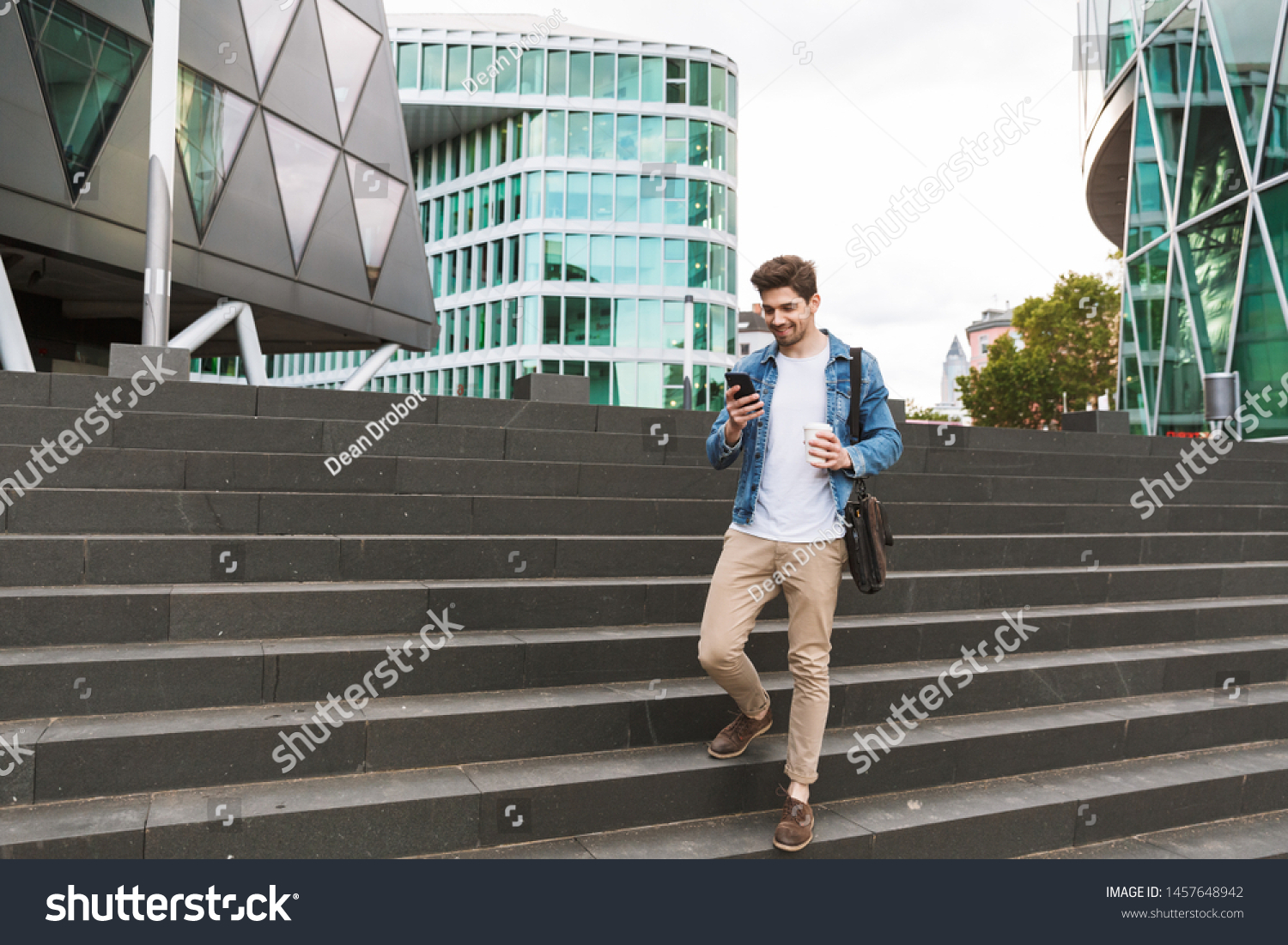 Handsome smiling young man dressed casually spending time outdoors at the city, using mobile phone while walking down stairs #1457648942