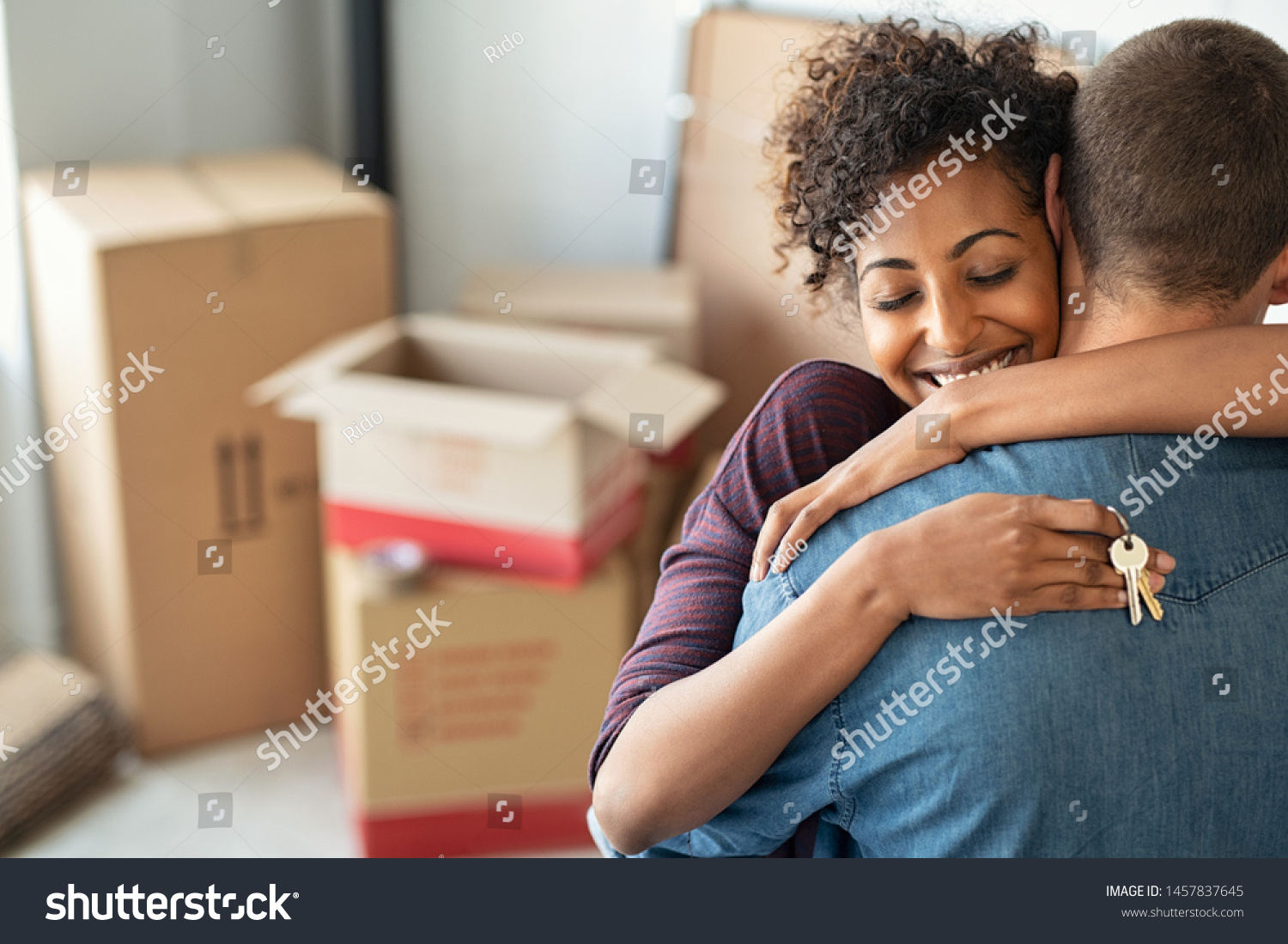 Young african woman holding home keys while hugging boyfriend in their new apartment after buying real estate. Lovely girl holding keys from new home and embracing man. Couple around cardboard boxes. #1457837645