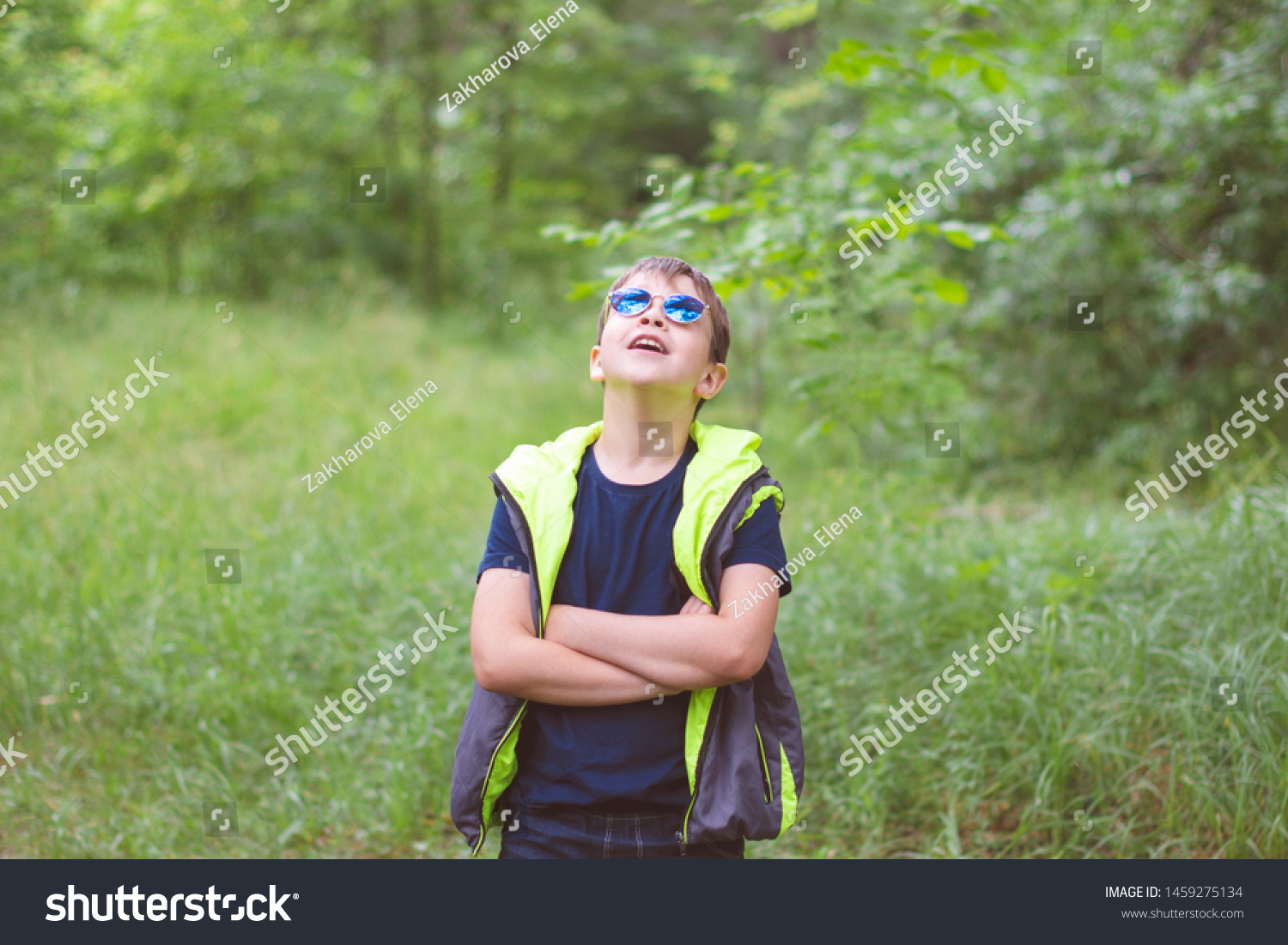 Trendy boy in a trendy blue sunglasses and a bright vest in the summer on a background of green nature. #1459275134