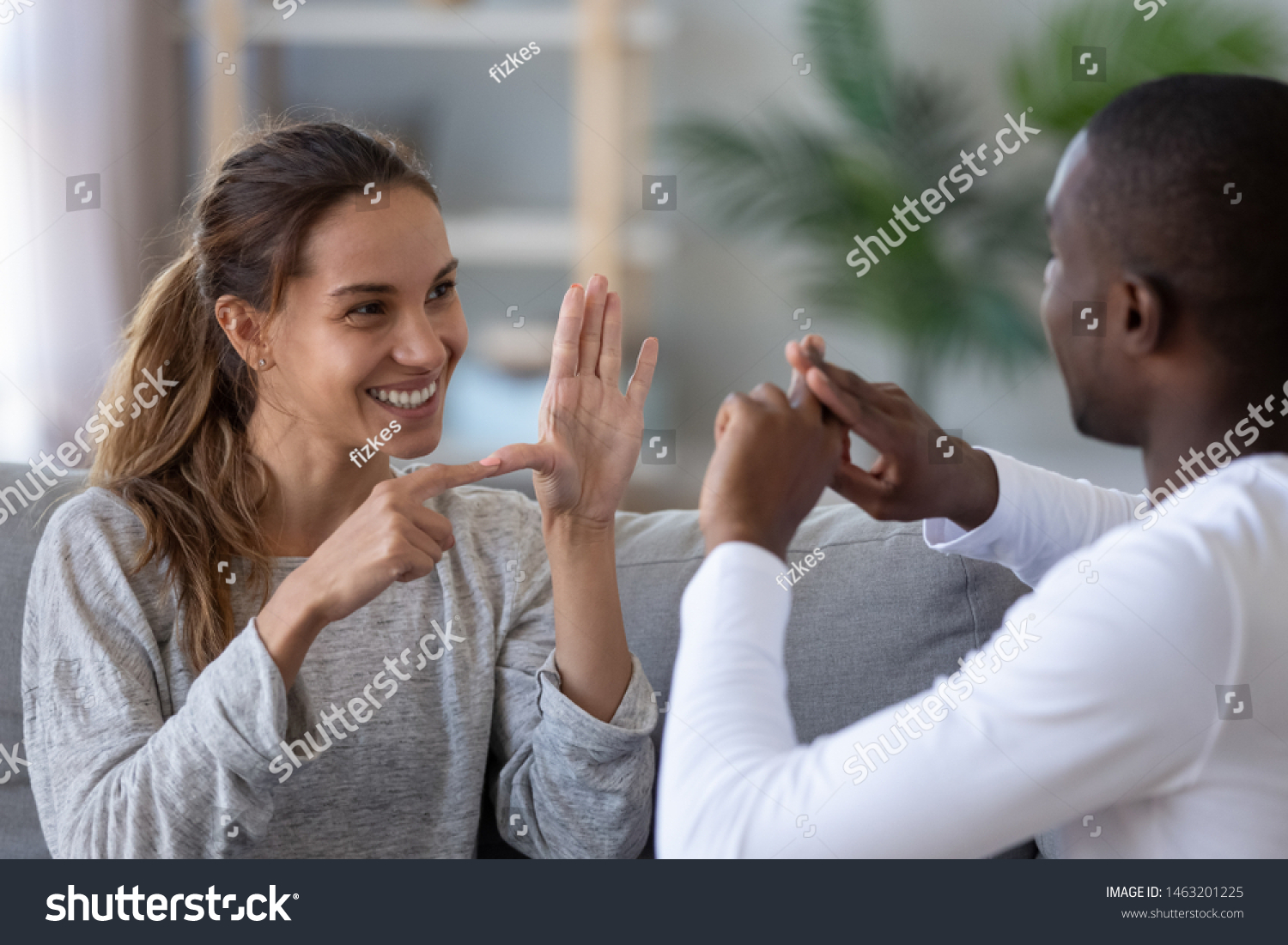 Smiling mixed ethnicity couple or interracial friends talking with sign finger hand language, happy two deaf and mute hearing impaired people communicating at home sit on sofa showing hand gestures #1463201225