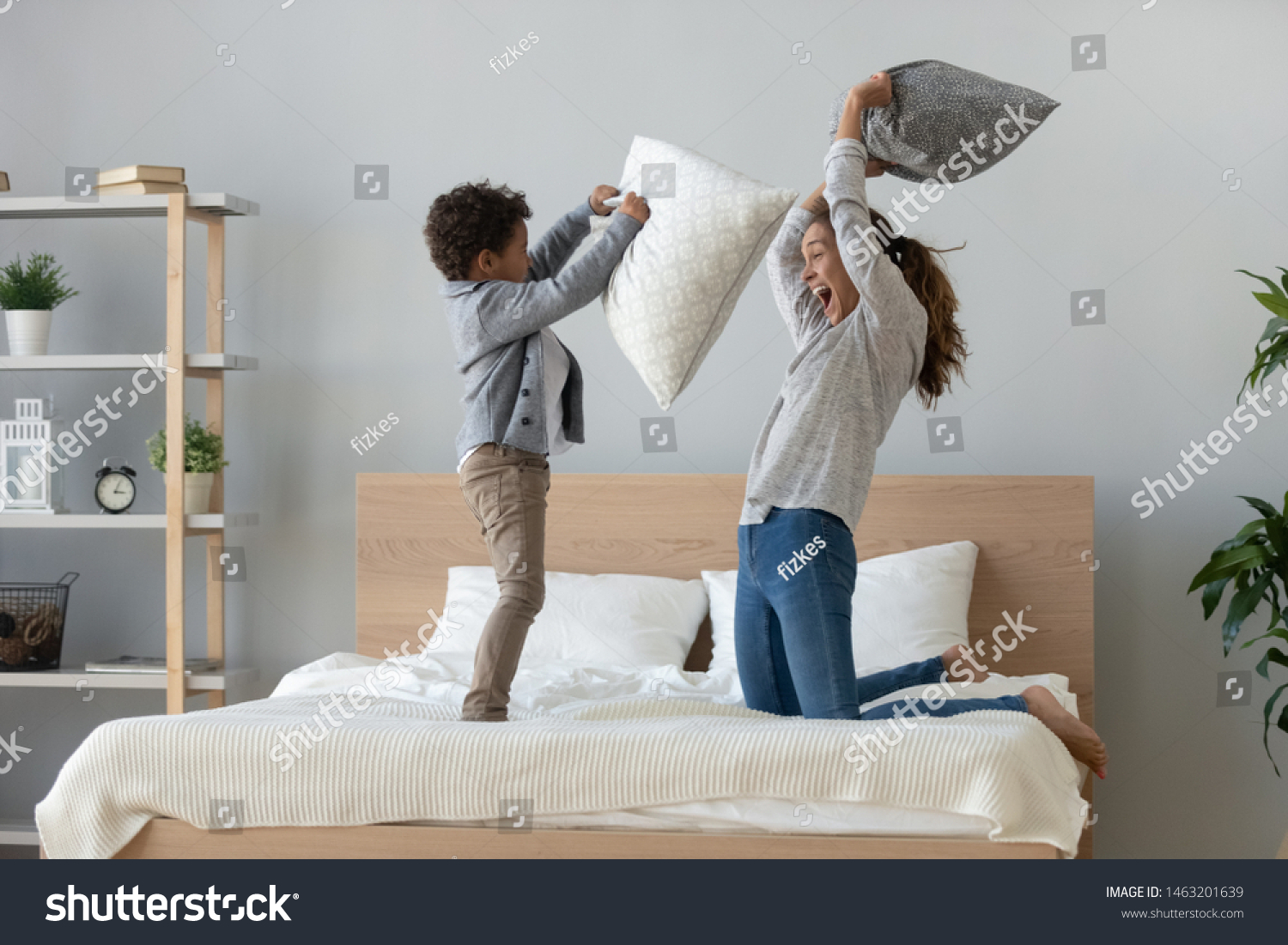 Funny happy african mixed race ethnicity family mum and little cute son having fun pillow fight on bed, young mother laughing playing funny game enjoy leisure activity with small child boy in bedroom #1463201639