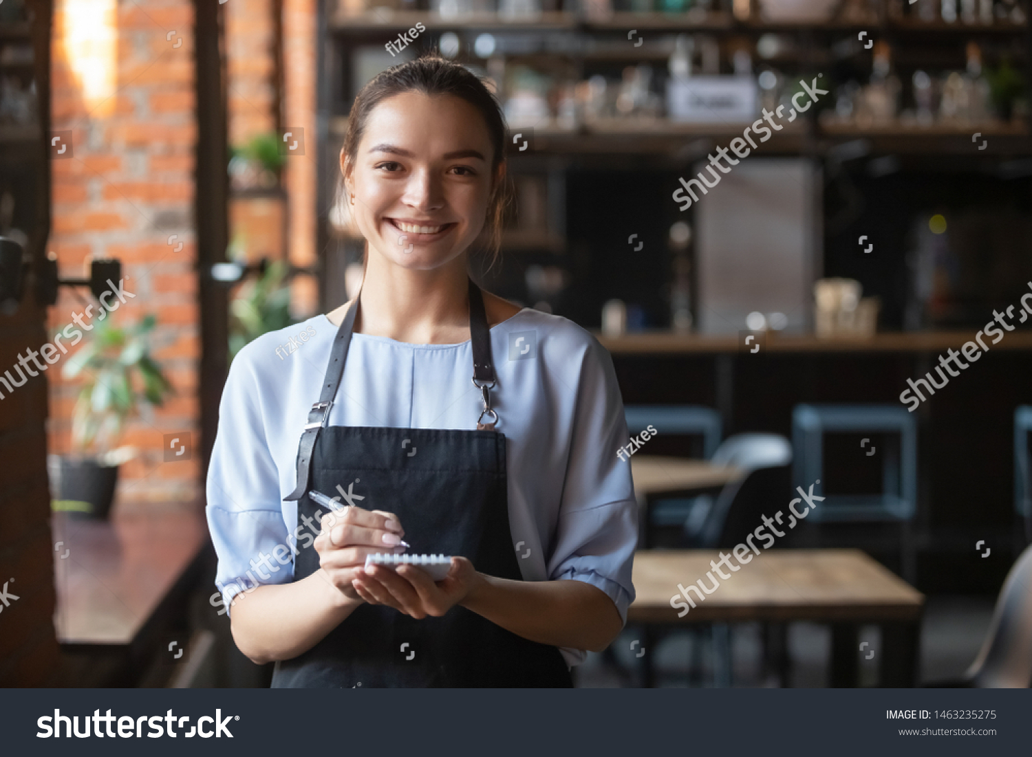 Portrait of smiling millennial waitress standing wearing uniform holding notebook looking at camera, positive happy cafe or restaurant female staff in apron ready to take order. Good service concept #1463235275