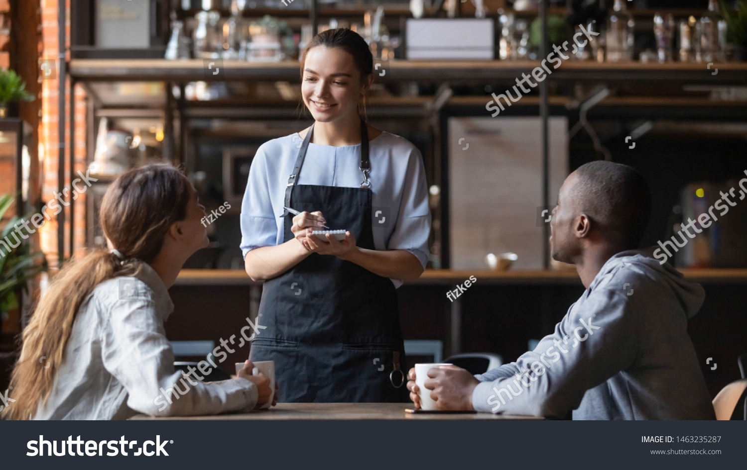 Smiling millennial waitress with notebook taking order from young multiracial client couple, diverse friends relax hang out in cafe or restaurant speak with staff enjoying good service and atmosphere #1463235287