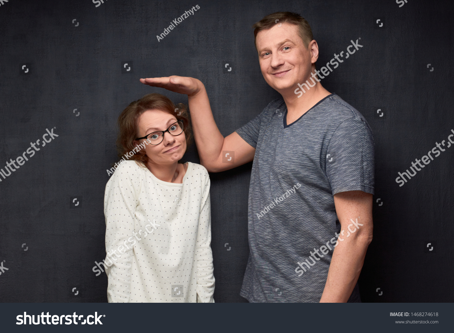 Studio waist-up shot of tall man smiling and showing with hand at height of short girl standing beside him and looking with perplexity at camera, over gray background. Variety of person's heights #1468274618