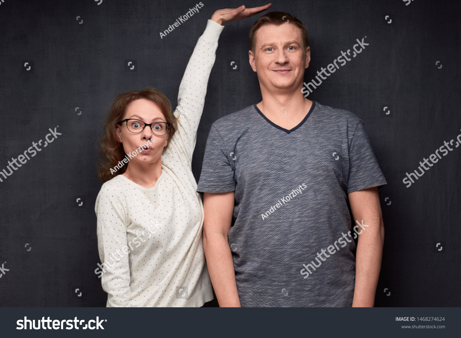 Studio waist-up shot of amazed short woman pulling up and showing with hand at height of tall man standing beside her, smiling and looking at camera, over gray background. Variety of person's heights #1468274624