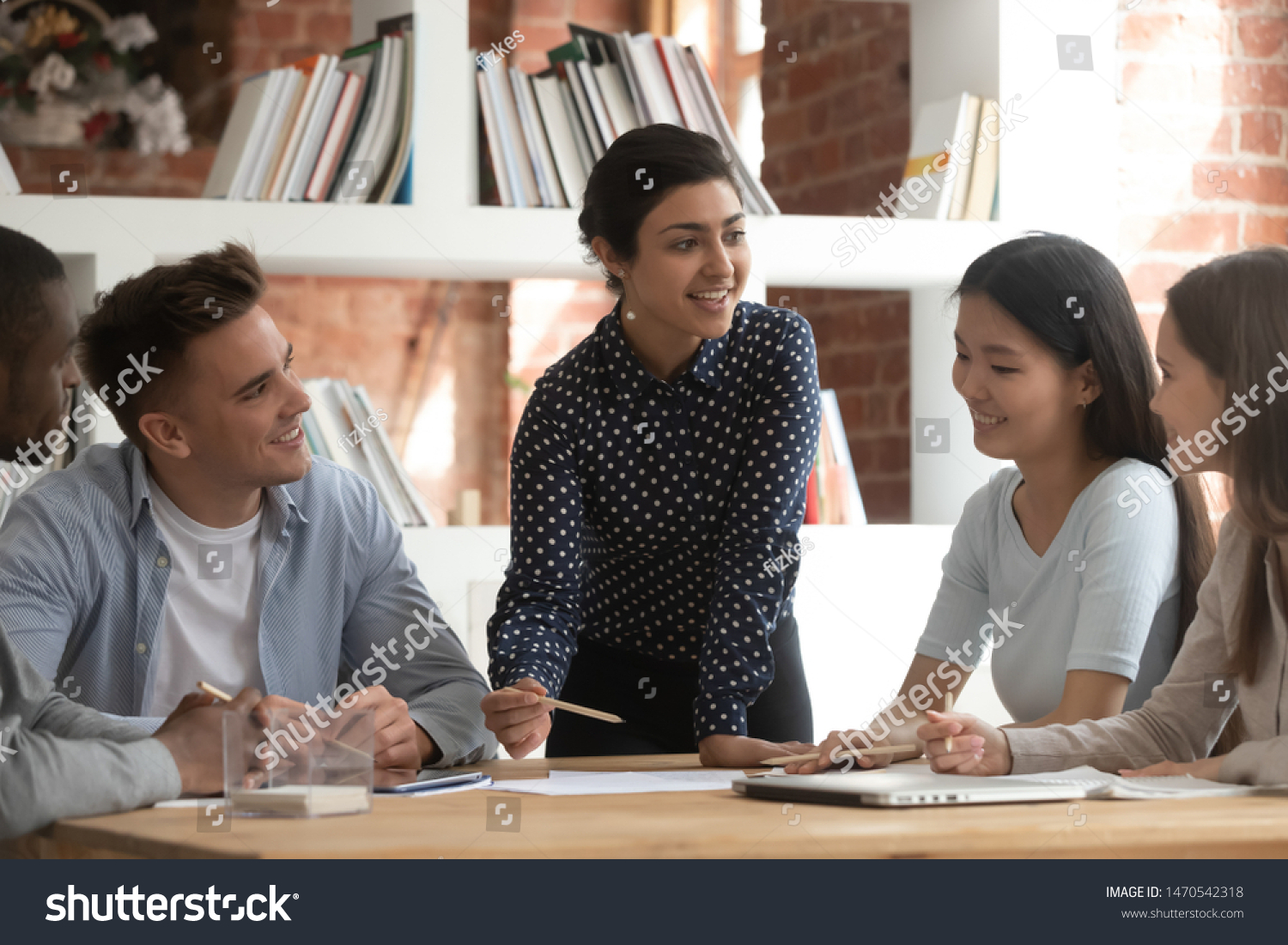 Cheerful indian female team leader involved in studying process, explaining new material to mixed race friends. Group of smiling diverse students discussing together school project ideas in classroom. #1470542318