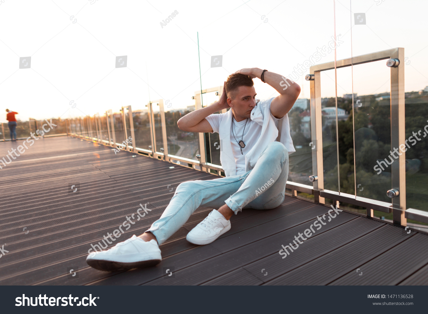 Young urban man with a stylish hairstyle in a stylish T-shirt in vintage jeans in white sneakers posing while sitting on a wooden floor on the terrace. Attractive guy model is enjoying the sunset.  #1471136528