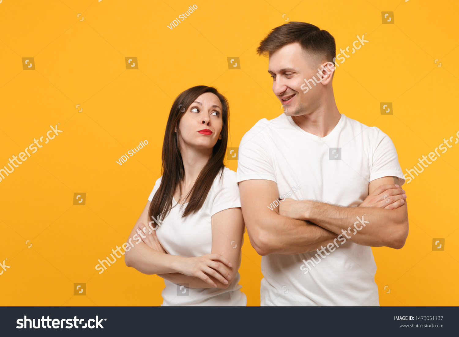 Smirked young couple two friends guy girls in white blank empty design t-shirts posing isolated on yellow orange wall background. People lifestyle concept. Mock up copy space. Holding hands crossed #1473051137