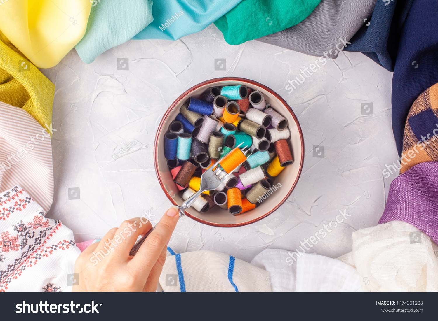 Woman holds fork with spool of thread under the bowl with spools in fabric multi-colored frame on grey background #1474351208