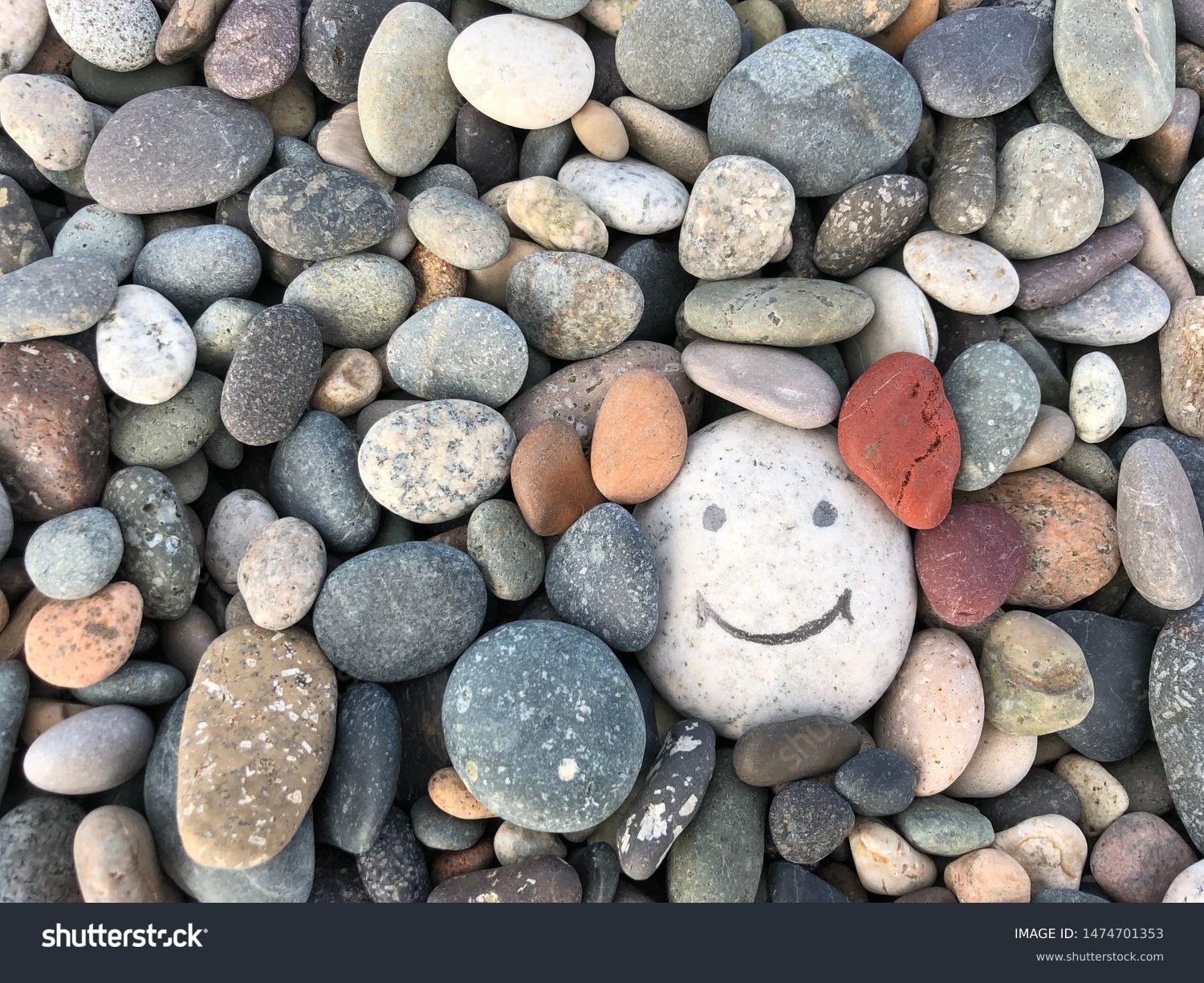 Stone with a painted smile. On the shore, one stone stands out from the others. On a small stone is an image of a happy face. Concept: joy, happiness, positive, kindness. #1474701353