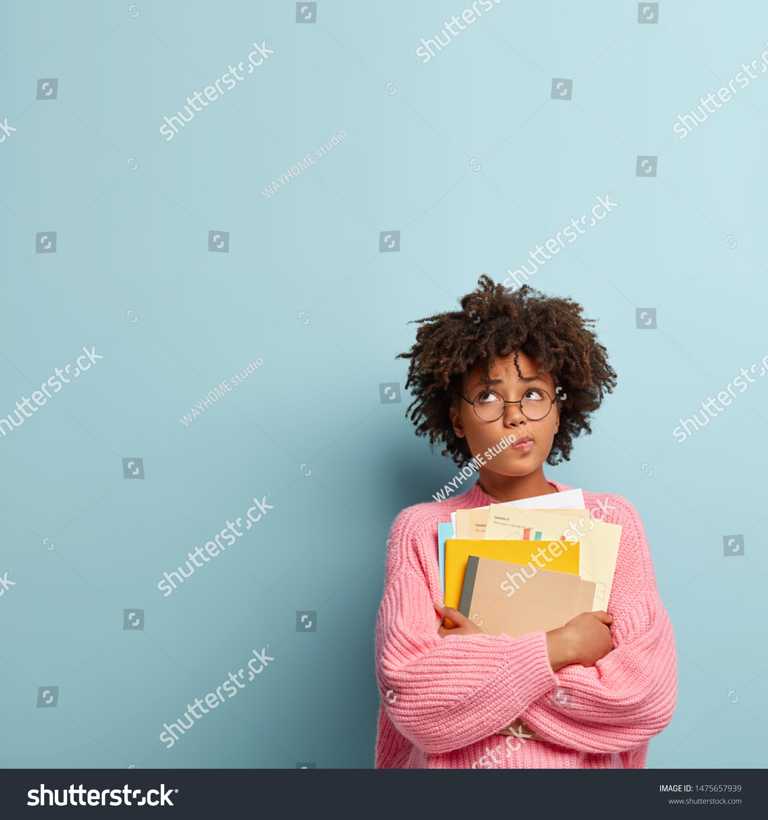 Pensive dark skinned college student holds papers and textbooks, purses lips and focused upwards, wears rosy knitted sweater, stands against blue background with copy space for your advertisement #1475657939