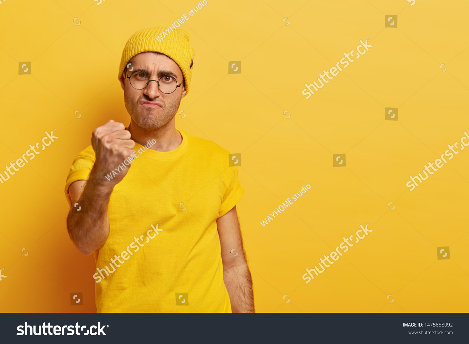 Serious powerful man needs respect, raises clenched fist, looks in dissatisfaction, doesnt afford being insulted, frowns in displeasure, feels annoyed and bothered, wears yellow hat, t shirt, eyewear #1475658092