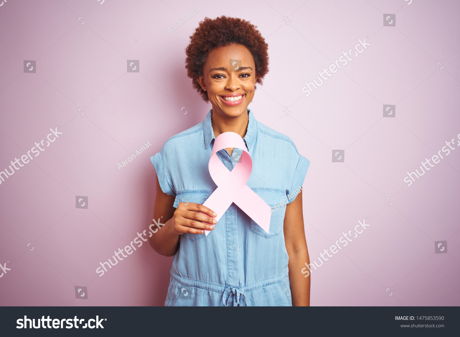 Young african american woman holding brest cancer ribbon over isolated pink background with a happy face standing and smiling with a confident smile showing teeth #1475853590