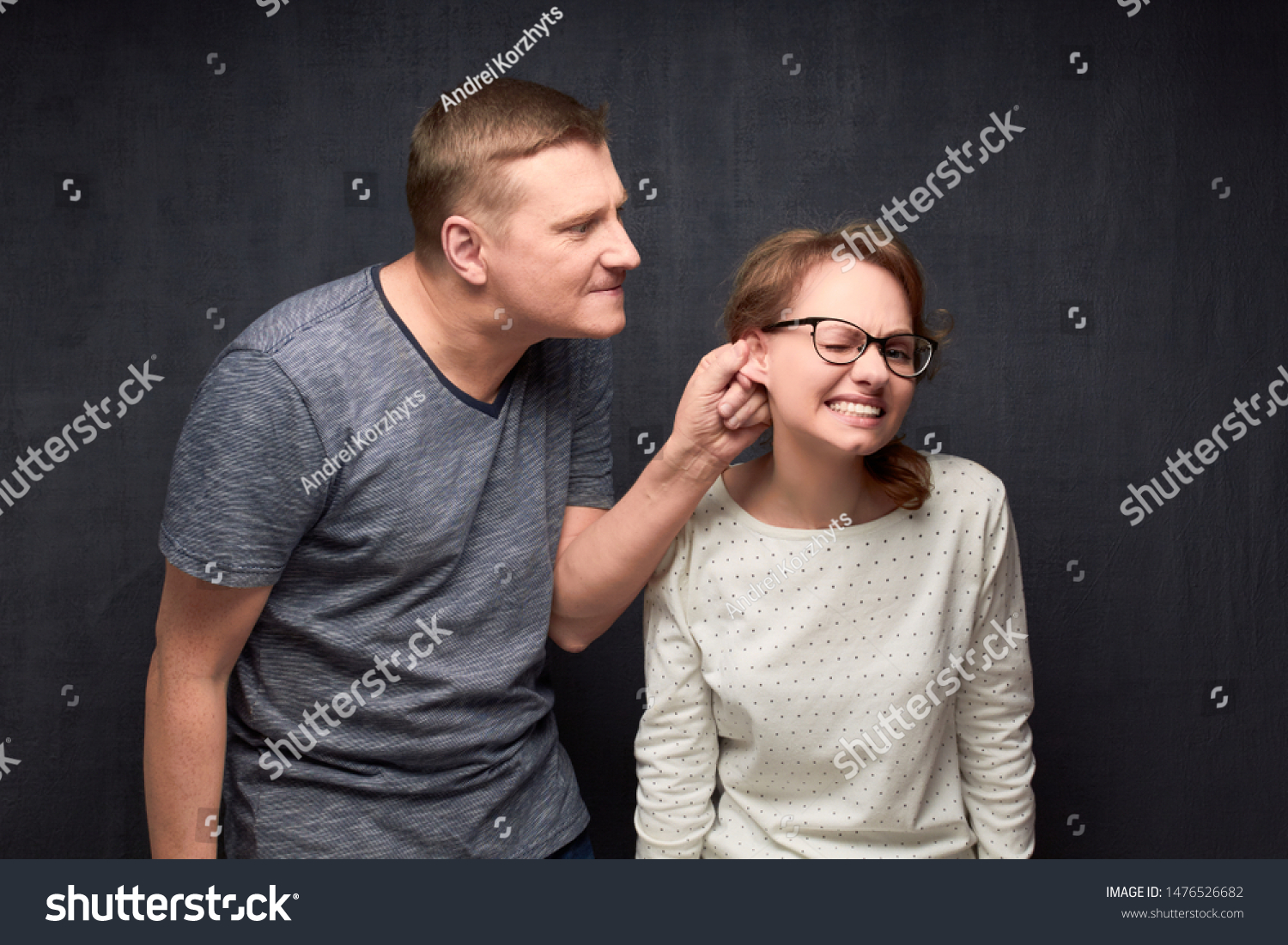 Studio waist-up shot of dissatisfied man frowning face, pulling woman by ear while scolding, woman is grimacing from pain, over gray background. Strict boyfriend and misbehaving girlfriend #1476526682