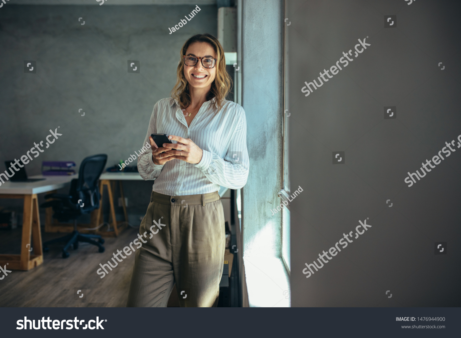 Smiling woman in casuals standing in office. Businesswoman with mobile phone in hand looking at camera. #1476944900