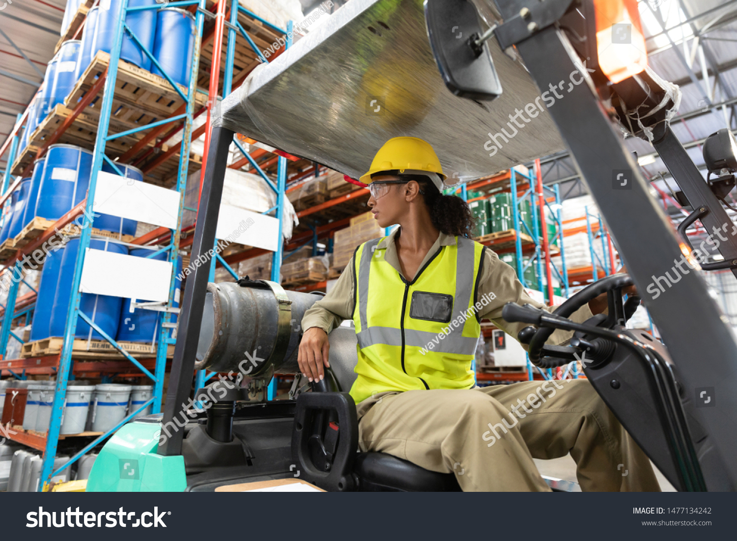 Low angle view of female staff driving forklift in warehouse. This is a freight transportation and distribution warehouse. Industrial and industrial workers concept #1477134242