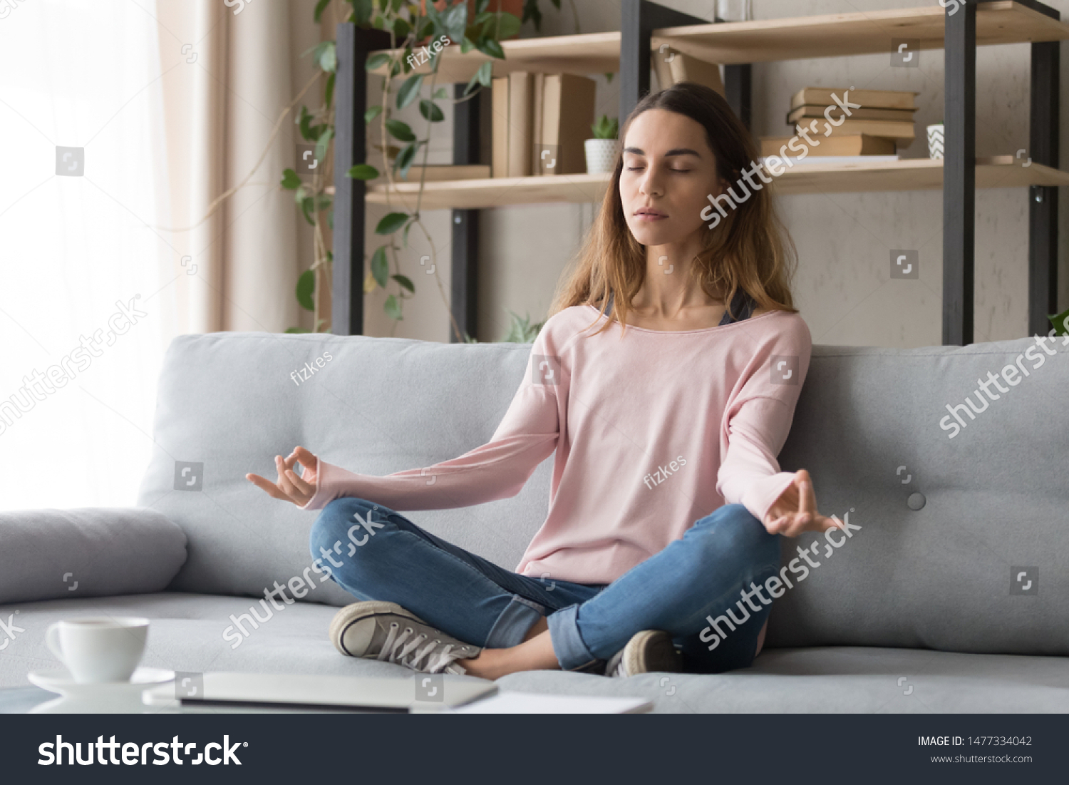 Carefree calm millennial freelance girl sitting on couch in lotus position and mudra gesture, relaxing, doing yoga practice. Young serene woman meditating, visualizing, healthy lifestyle concept. #1477334042