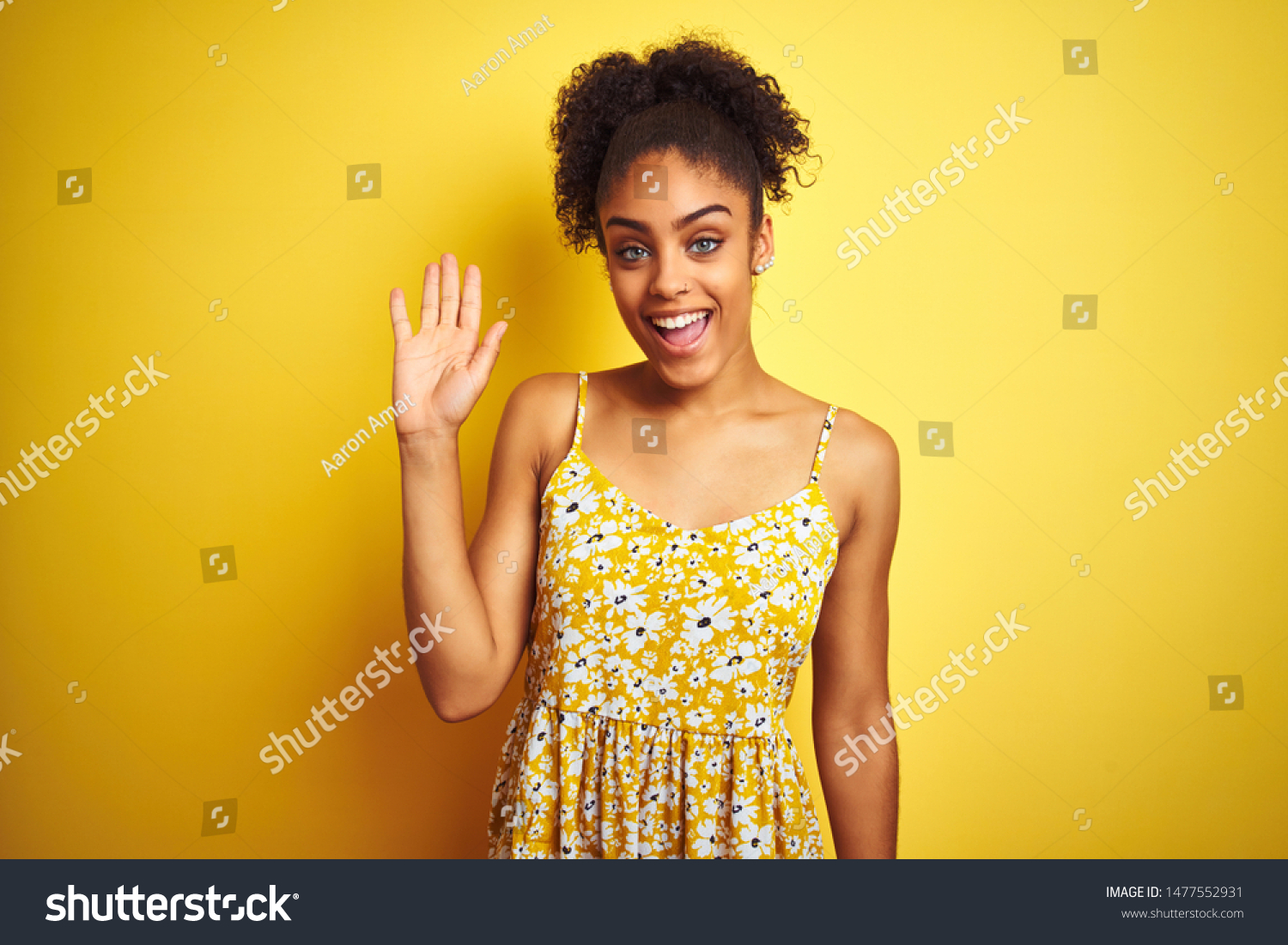 African american woman wearing casual floral dress standing over isolated yellow background Waiving saying hello happy and smiling, friendly welcome gesture #1477552931