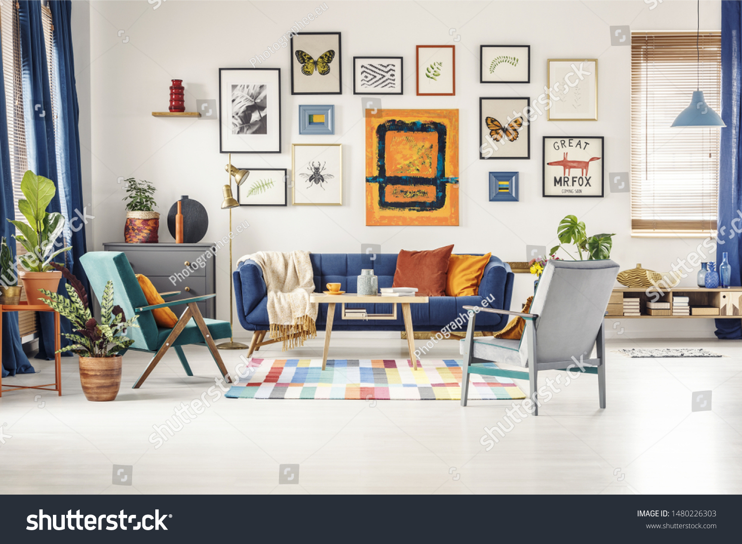 Simple posters gallery hanging on the wall in bright living room interior with blue sofa, two armchairs, fresh plants and wooden coffee table standing on colorful carpet #1480226303
