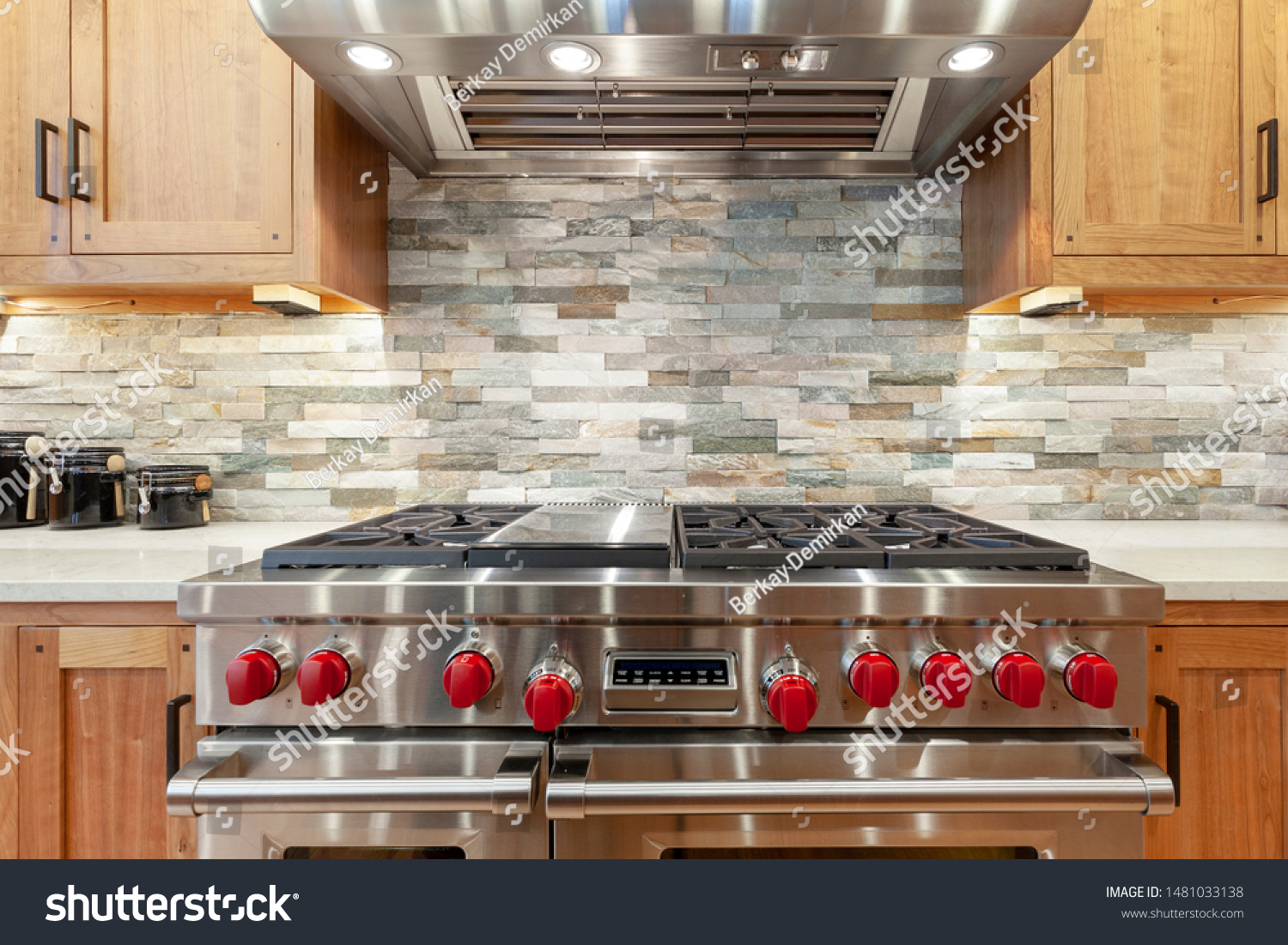 Stainless steel oven and stove with red knobs, stainless steel hood, brown kitchen cabinets, slate mosaic backsplash, natural stone, black jars #1481033138