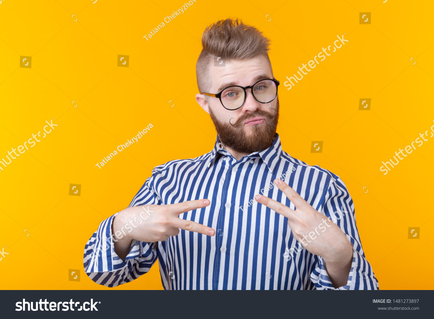 Charming confident young fashion hipster man with glasses and a beard shows victory gesture posing over a yellow background. The concept of self-confidence and funny. #1481273897