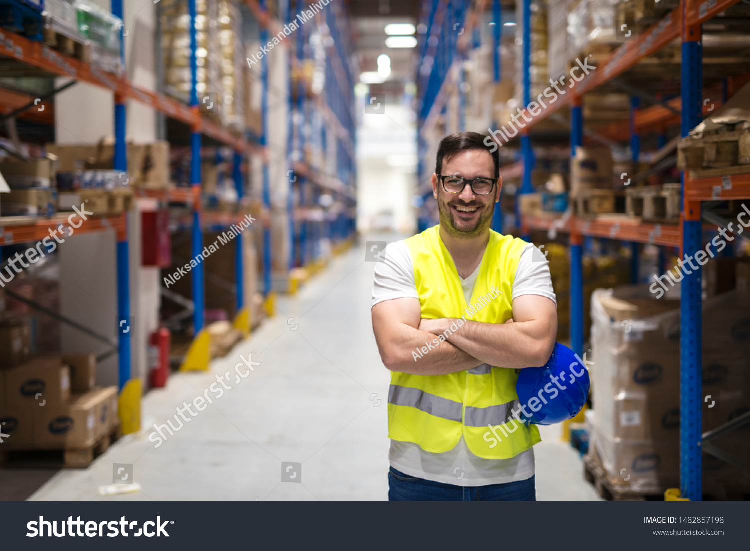 Portrait of middle aged caucasian warehouse worker standing in large warehouse distribution center with arms crossed. In background shelves with goods. Worker smilling and looking to the camera. #1482857198