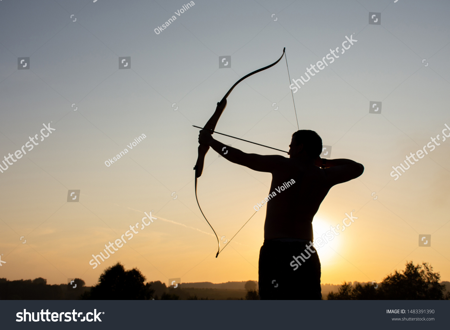 Silhouette of a handsome man with an ancient weapon bow and arrow on a background of sky and sunset #1483391390
