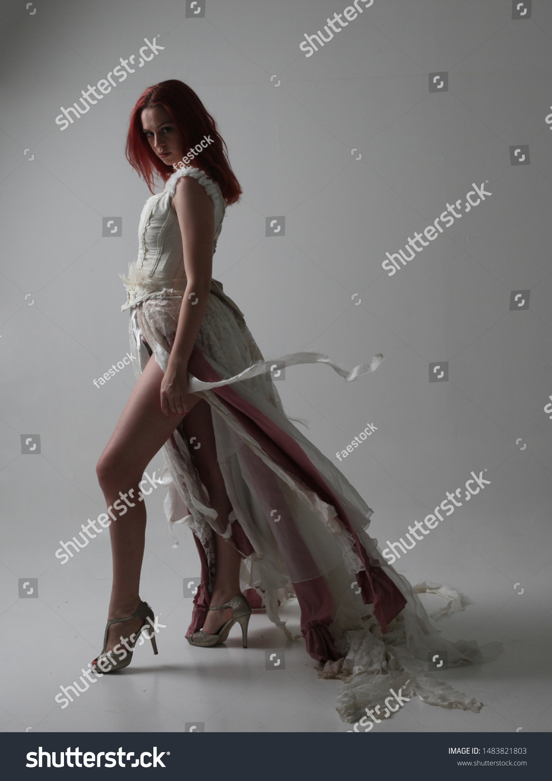 full length portrait of red haired girl wearing torn and tattered wedding dress. Standing pose against a studio background with contrasty shadow lighting. #1483821803