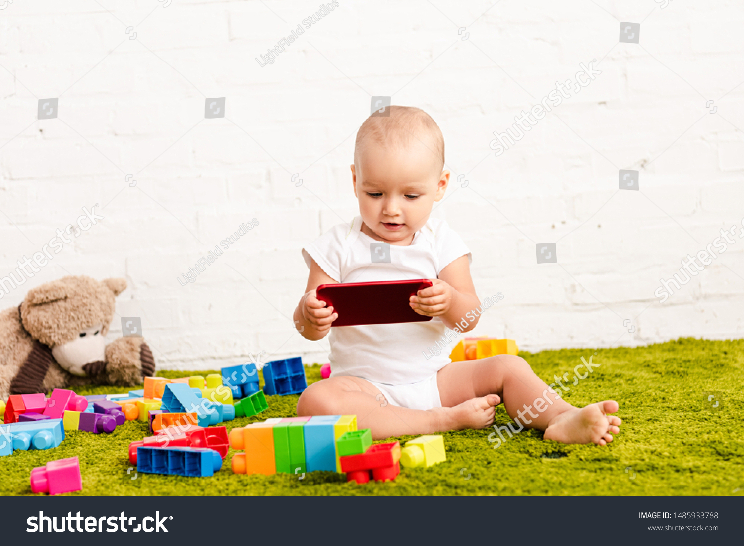 adorbale kid sitting among toys on green floor and holding digital device #1485933788