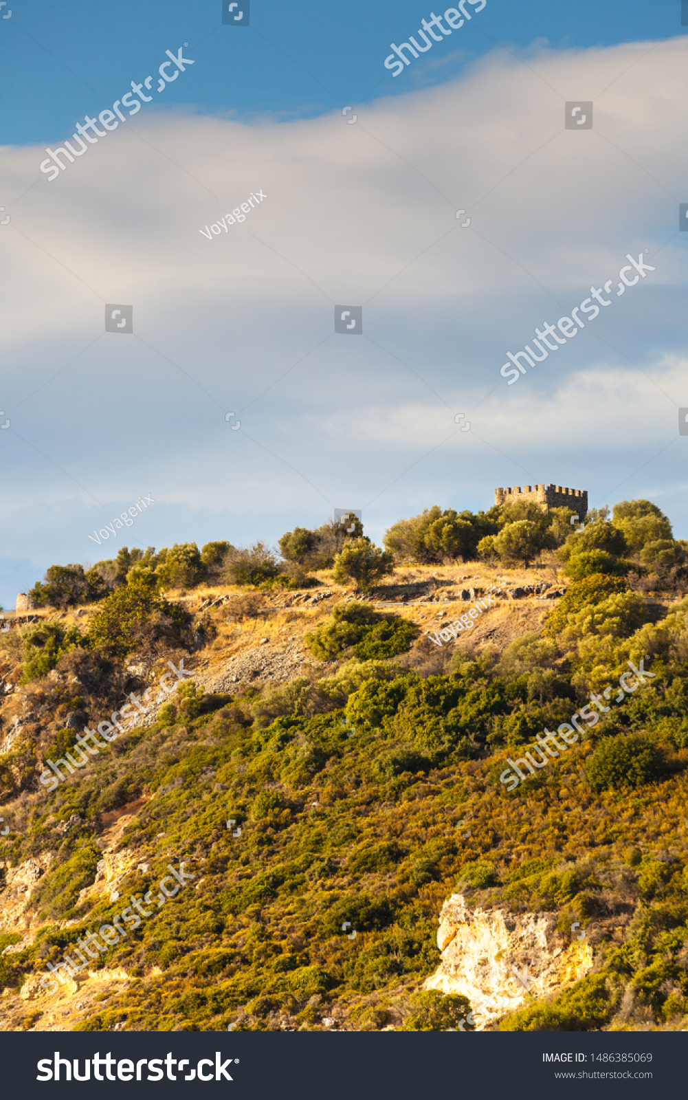 Greek idyllic hills with small shrubs of Mediterranean flora and little castle against cloudy sky. #1486385069