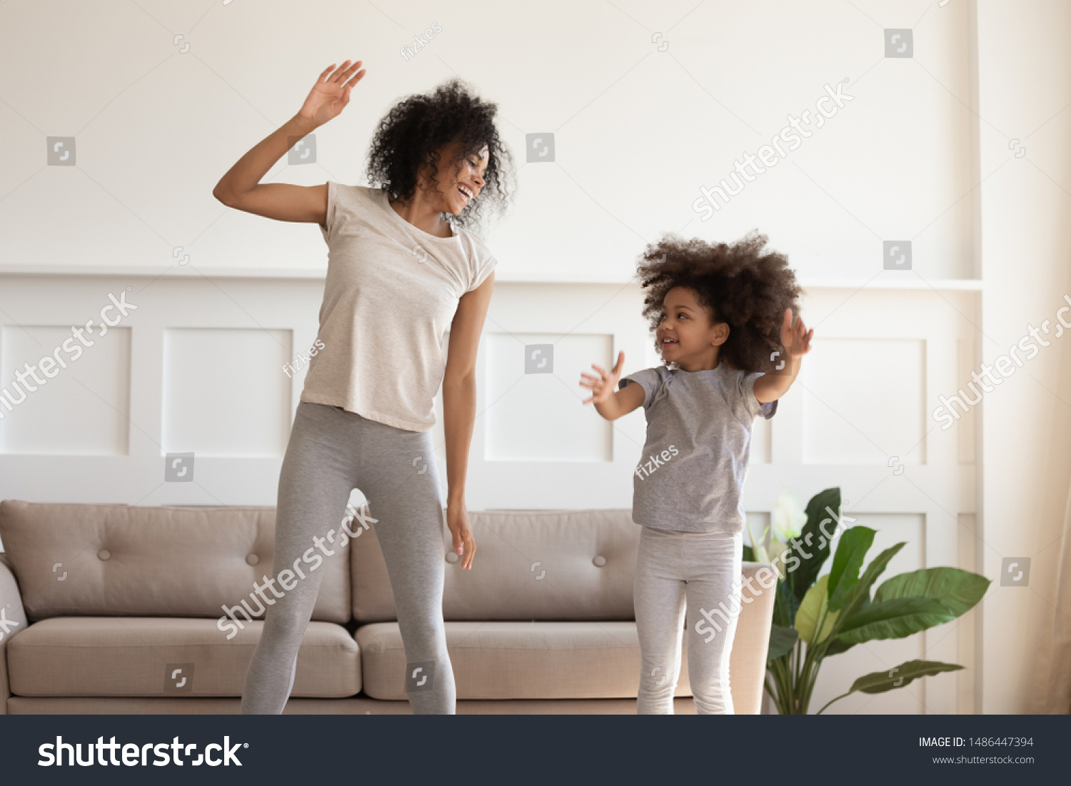Happy smiling african american mother doing morning exercises with excited little preschooler daughter in stylish living room at home. Overjoyed black family dancing, jumping, having fun together. #1486447394