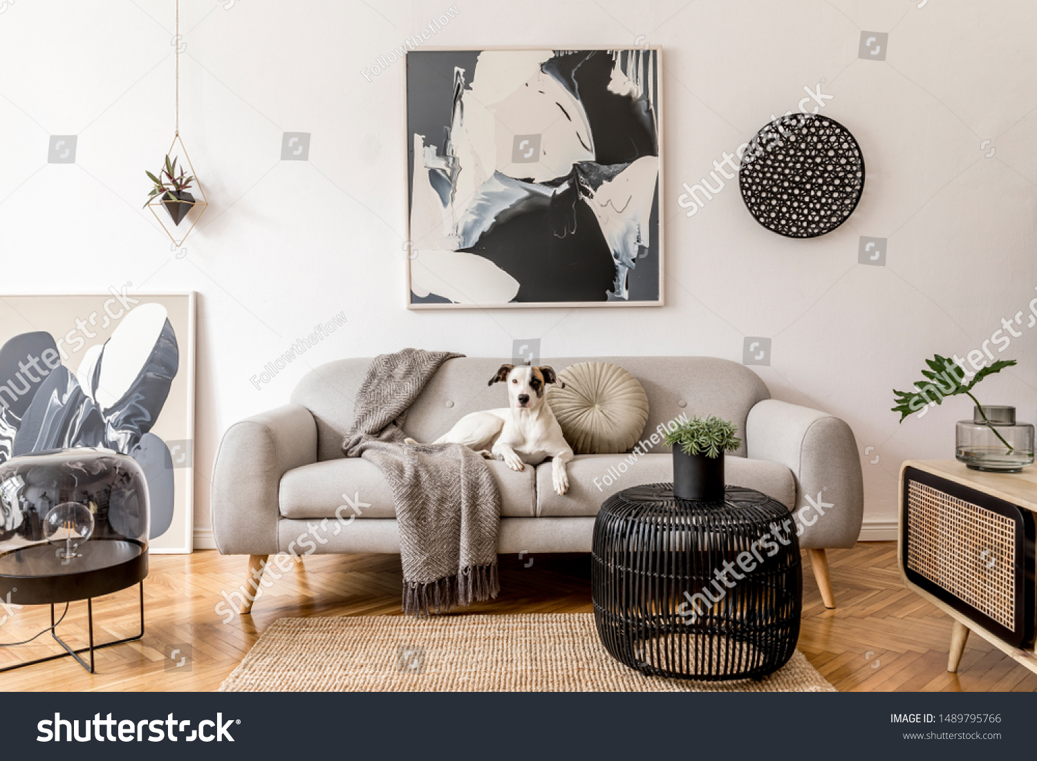 Stylish and scandinavian living room interior of modern apartment with gray sofa, design wooden commode, black table, lamp, abstract paintings on the wall. Beautiful dog lying on the couch. Home decor. #1489795766