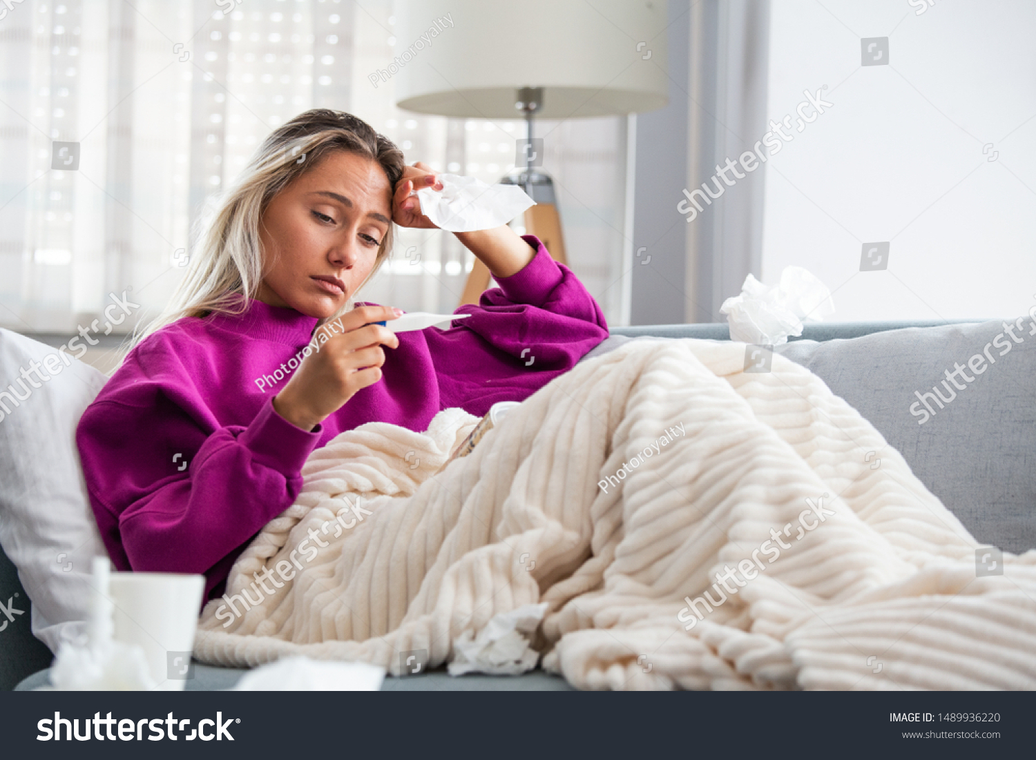 Sickness, seasonal virus problem concept. Woman being sick having flu lying on sofa looking at temperature on thermometer. Sick woman lying in bed with high fever. Cold flu and migraine. #1489936220