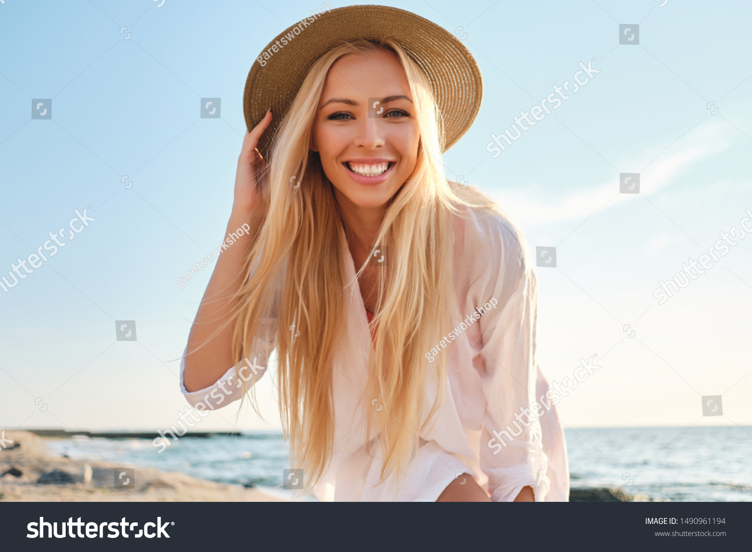 Young attractive smiling blond woman in white shirt and hat joyfully looking in camera with sea on background #1490961194
