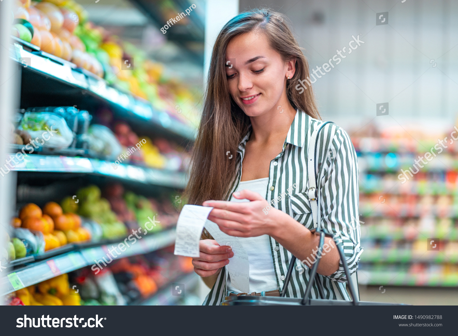 Young woman with shopping basket checks and examines a sales receipt after purchasing food in a grocery store. Customer buying products at supermarket  #1490982788